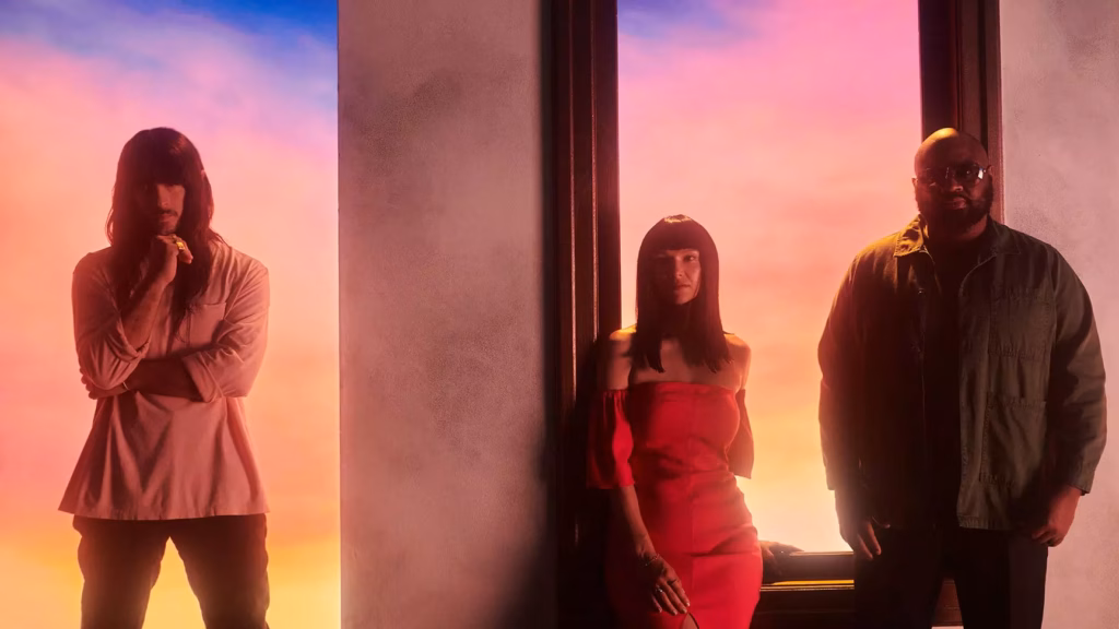 Khruangbin poses against a softly backlit pastel background featuring a sunset