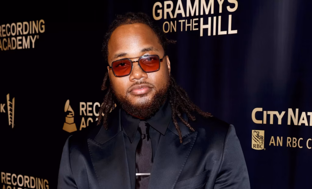 Leon Thomas at the 2024 Grammys On The Hill Awards. Thomas is wearing dark red sunglasses and an all-black suit and standing in front of the 2024 Grammys On The Hill step-and-repeat background.