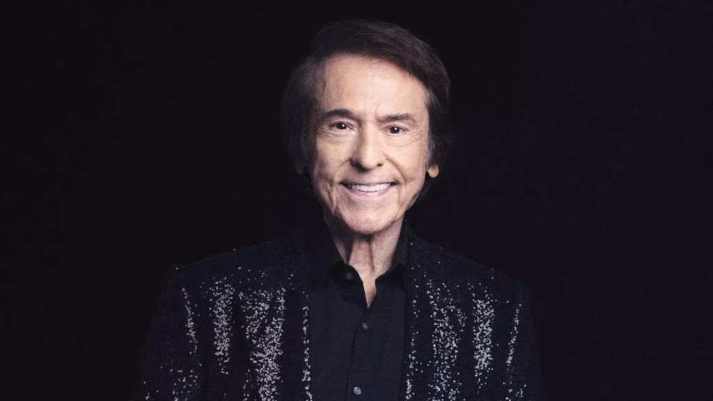 Photo of Spanish singer and 2025 Latin Recording Academy Person Of The Year honoree Raphael. He is smiling directly at the camera, wearing a black, sparkly jacket and a black dress shirt and set against a black background.