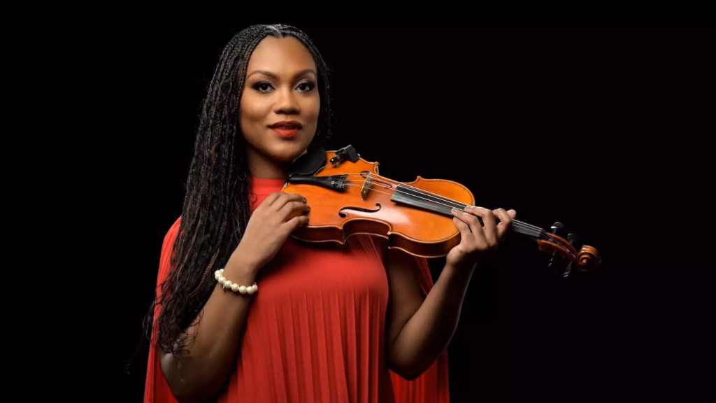 A press photo of Dr. Chelsey Green, Chair on the Board of Trustees of the Recording Academy. She is facing the camera, wearing a red dress and holding a violin in her hands.