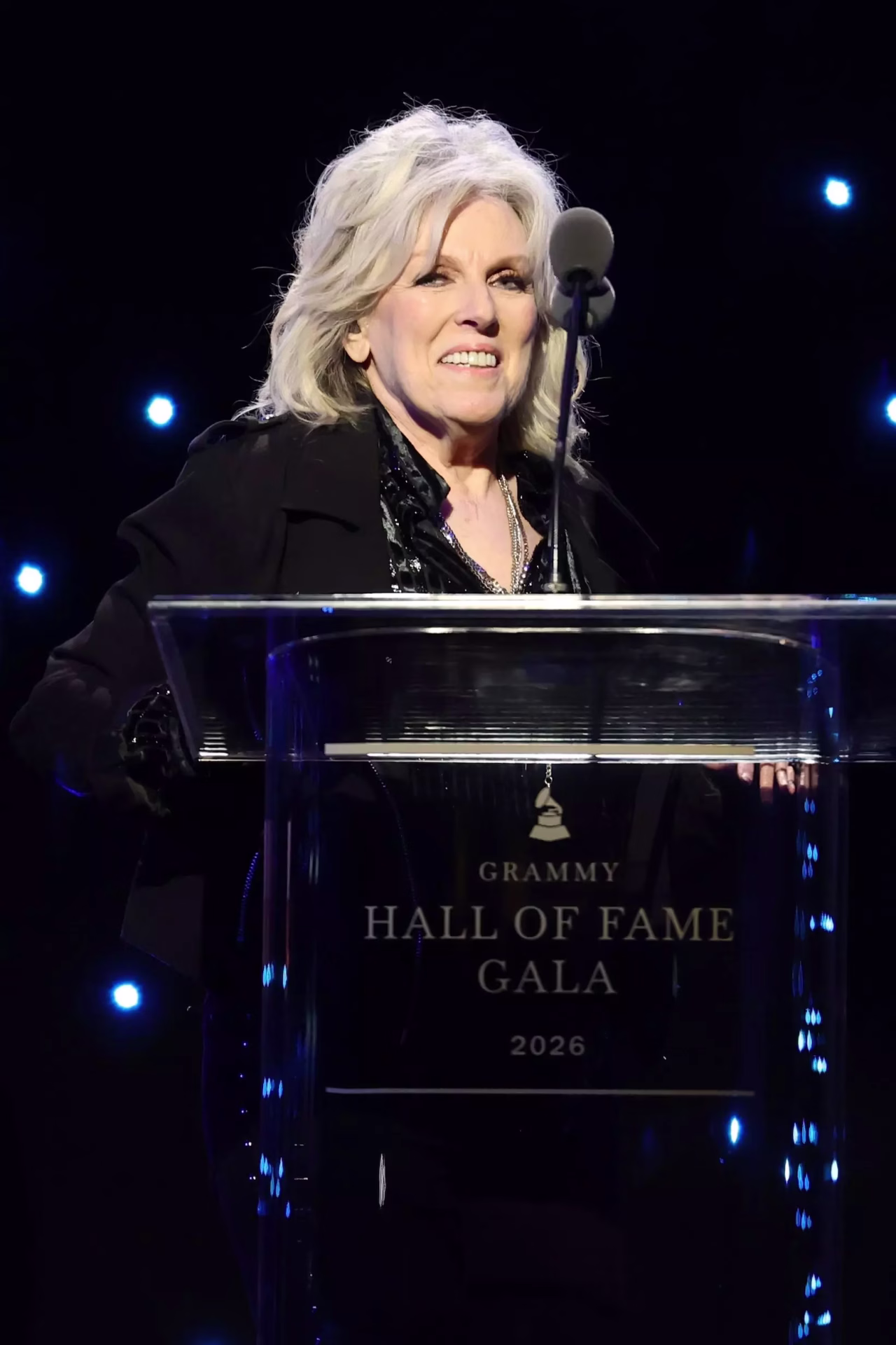 Lucinda Williams accepts an honor onstage during the 2026 Grammy Hall of Fame Gala on Friday, May 8, 2026, at The Beverly Hilton in Los Angeles. Lucinda Williams is wearing a black coat and black shirt and smiling in front of a microphone.