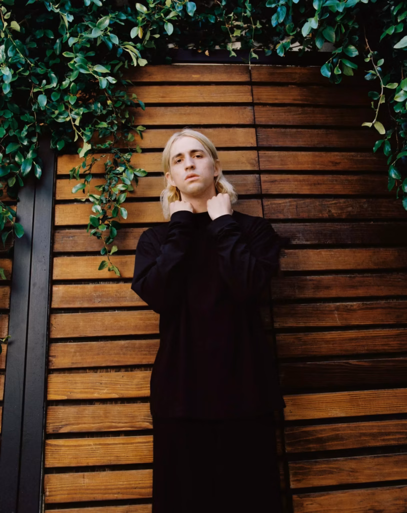 Porter Robinson poses in all-black, in front of a vine-covered fence